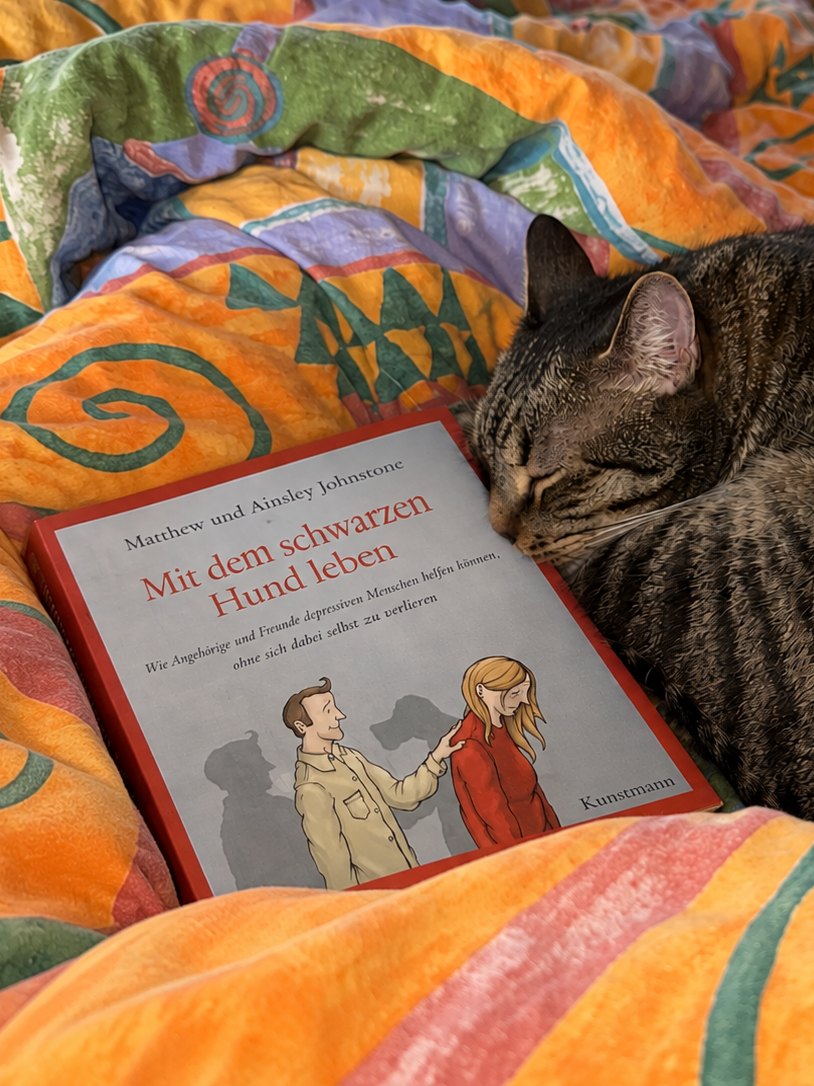 Das Buch „Mit dem schwarzen Hund leben“ liegt auf dem Bett, daneben schläft ein Kater